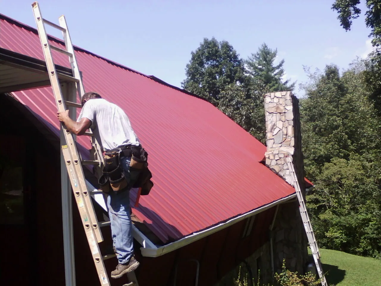 Roofer on a ladder beside a red metal roof and stone chimney — Soffit & Fascia Repair by Roofing Brothers in Mc Kinney